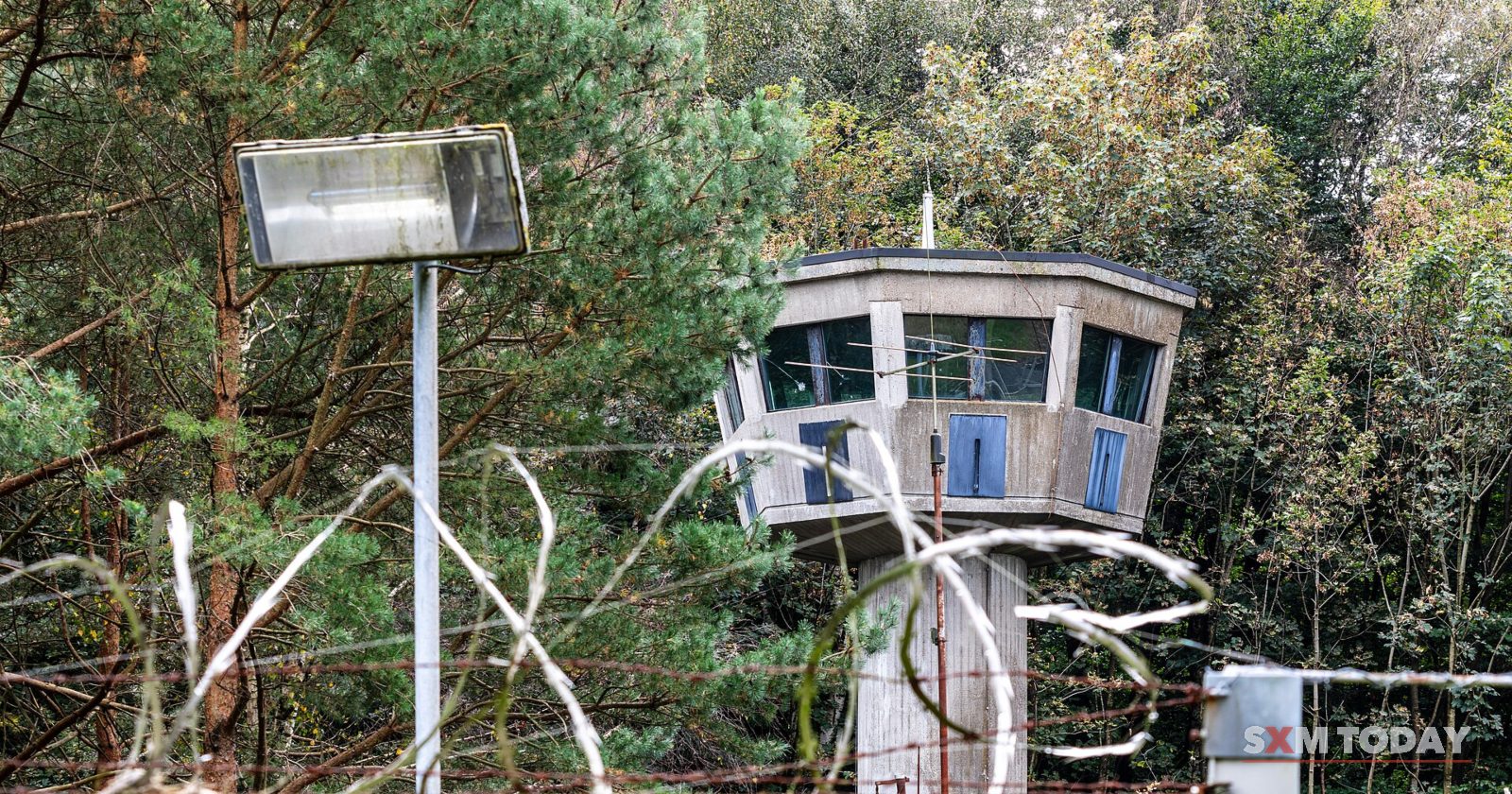 A guard tower with wide windows rises behind coils of razor wire and a floodlight on a fence.