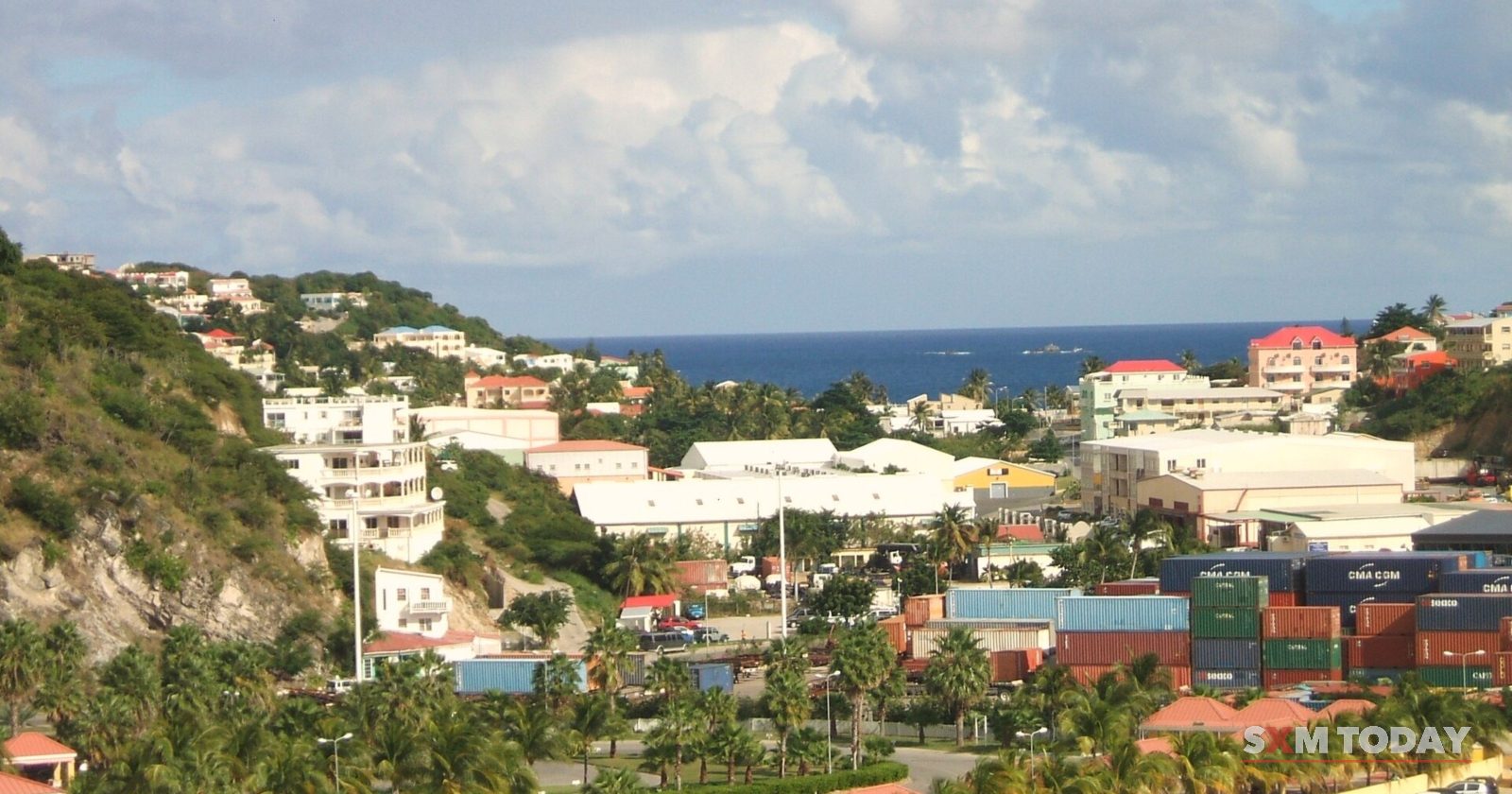 Houses climb a green hillside above palm trees, a car park and stacks of shipping containers, with the sea beyond.