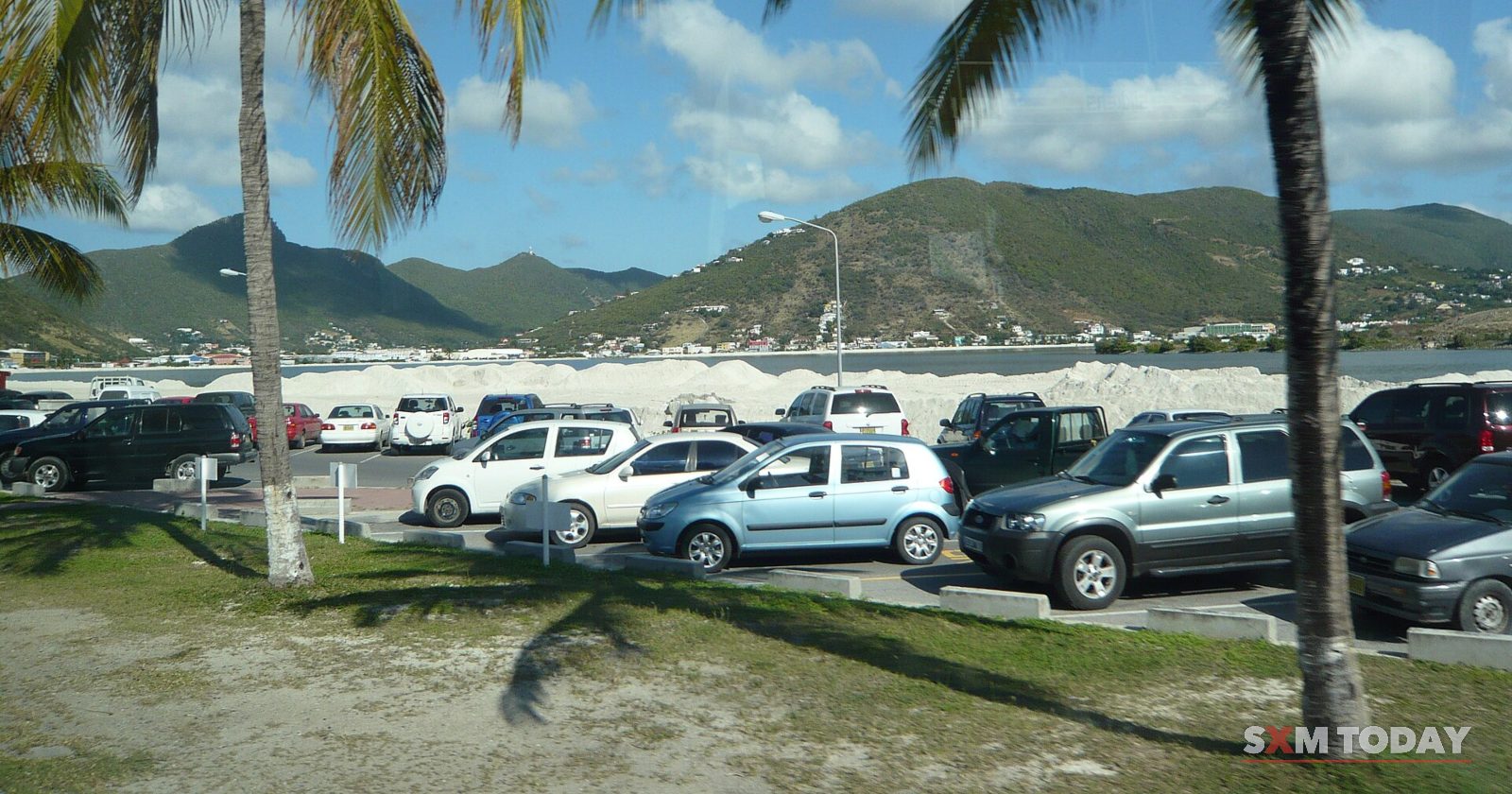 Rows of parked cars fill a car park shaded by tall palm trees, with green hills and a pale sky behind.