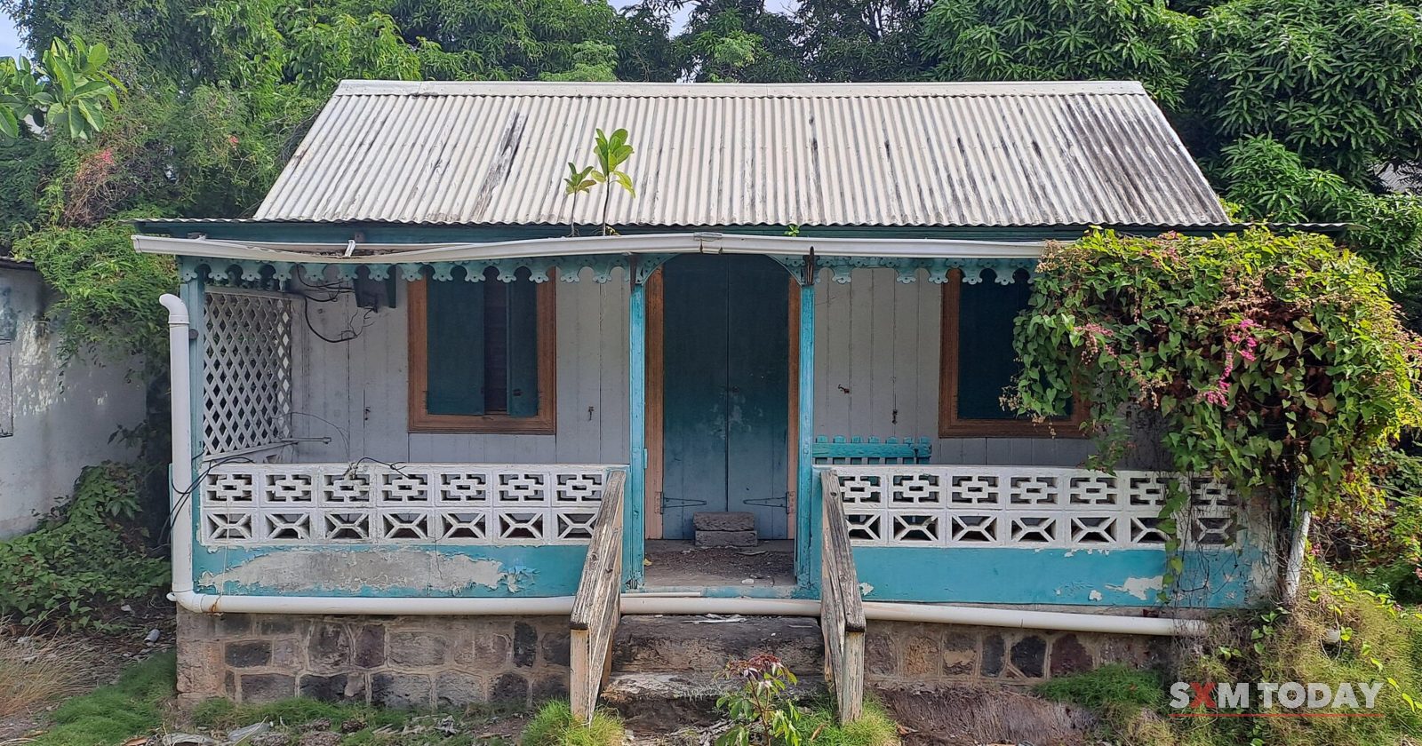A small weathered wooden house with a blue-trimmed veranda, patterned block railings and a corrugated roof stands on a stone base amid greenery.