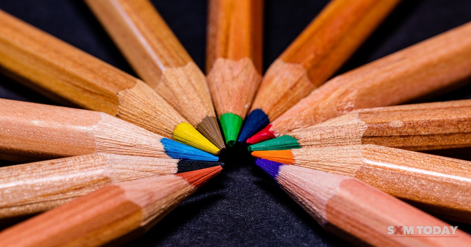 Sharpened wooden coloured pencils arranged in a star, their coloured tips meeting in the centre on a dark background.