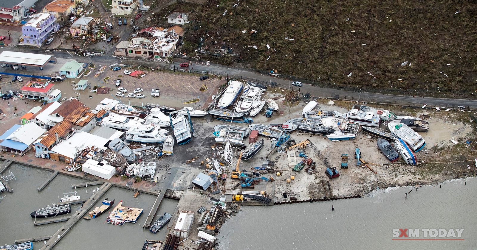 An aerial view of wrecked boats piled against a marina shore beside damaged buildings and stripped hillsides.