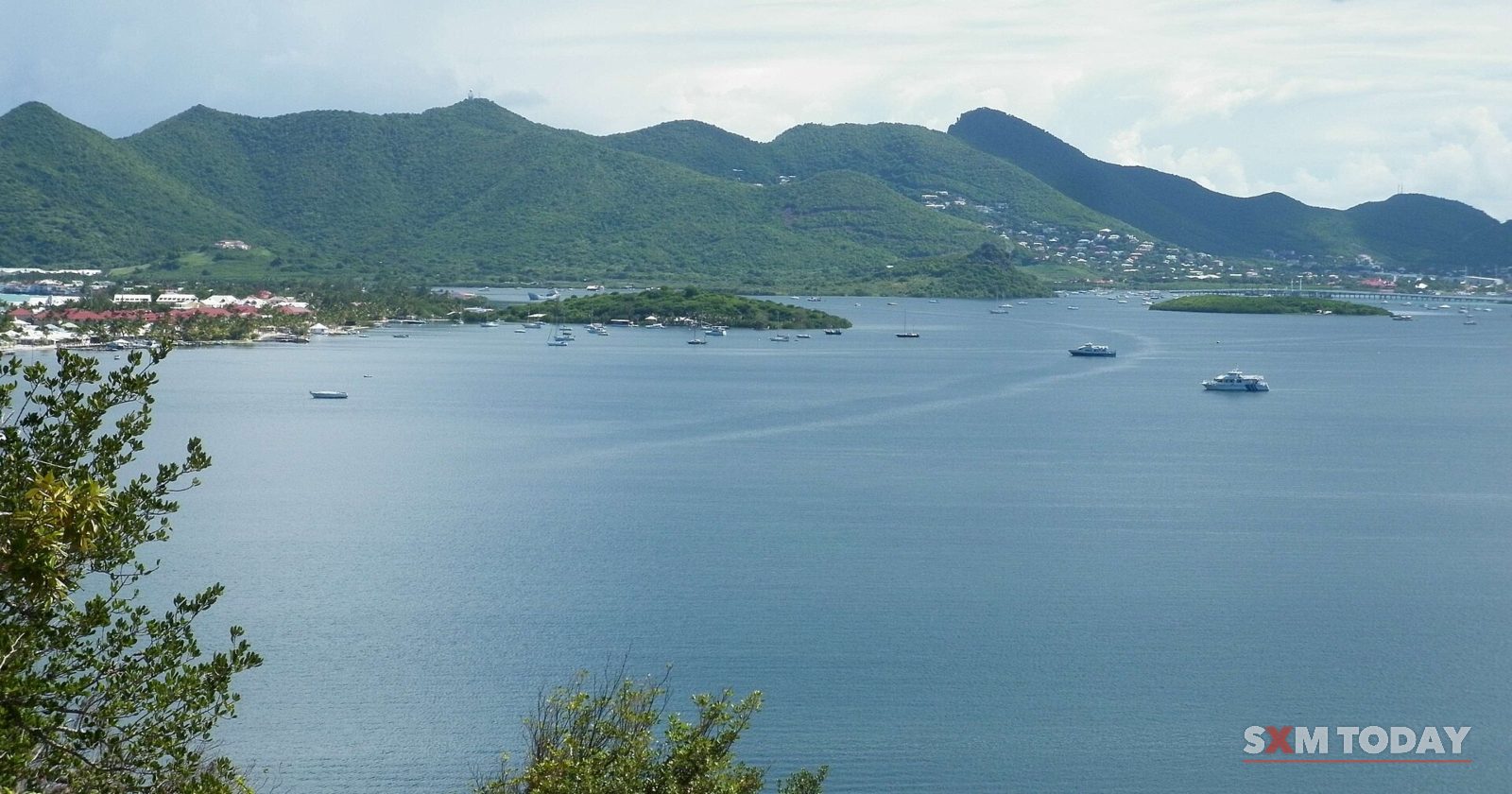 A wide calm lagoon with a few boats and small islands stretches toward green hills under a cloudy sky.