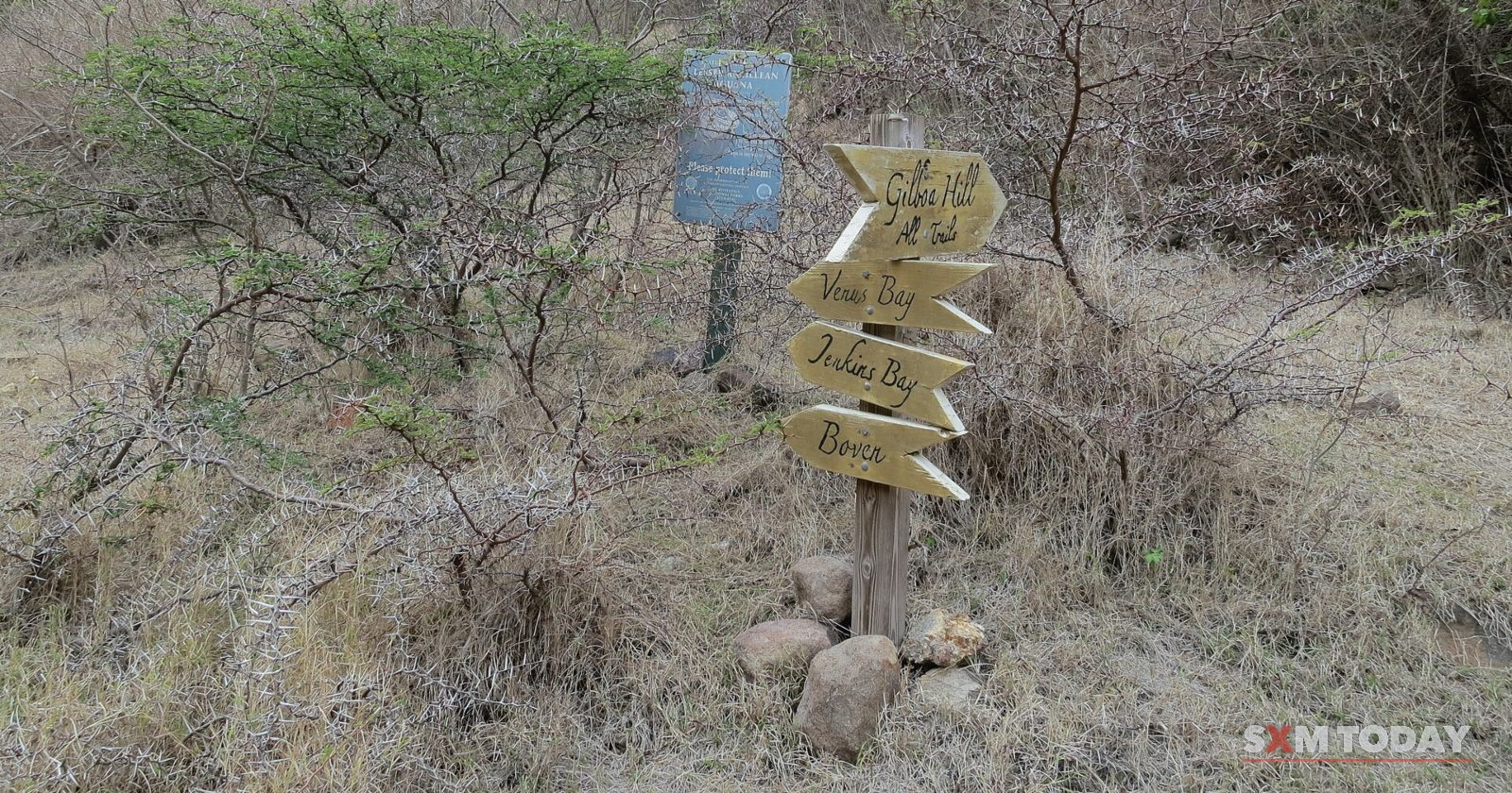 A wooden hiking signpost with arrow-shaped boards and an information panel stands in dry scrub.