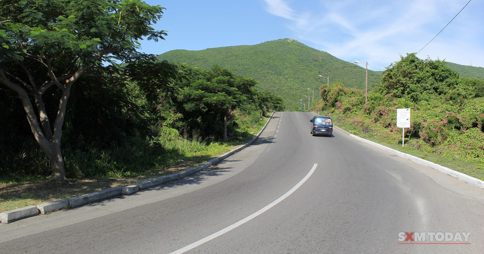 A two-lane road curves uphill between trees and green hills, with a single car approaching.