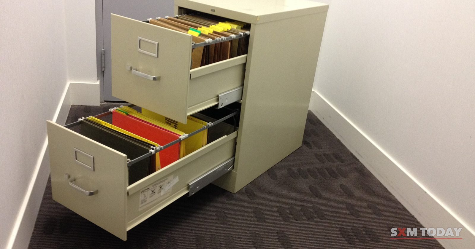 A beige two-drawer filing cabinet with both drawers pulled open, full of coloured folders, in the corner of a carpeted room.