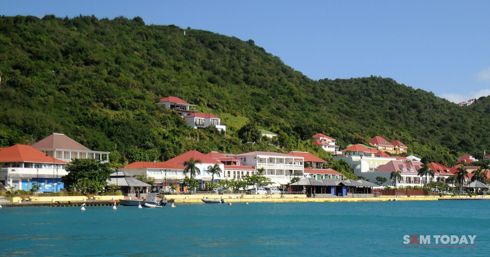 White and red-roofed buildings along a waterfront below a steep green hillside, seen across turquoise water.