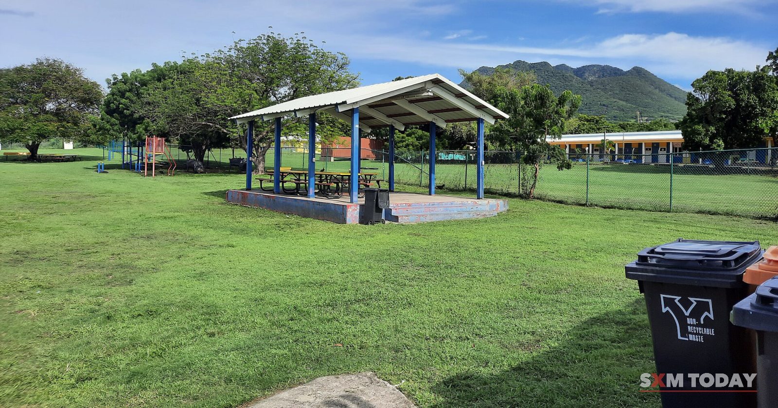 An open-sided shelter on a wide lawn with trees and green hills behind, and wheelie bins at the edge.