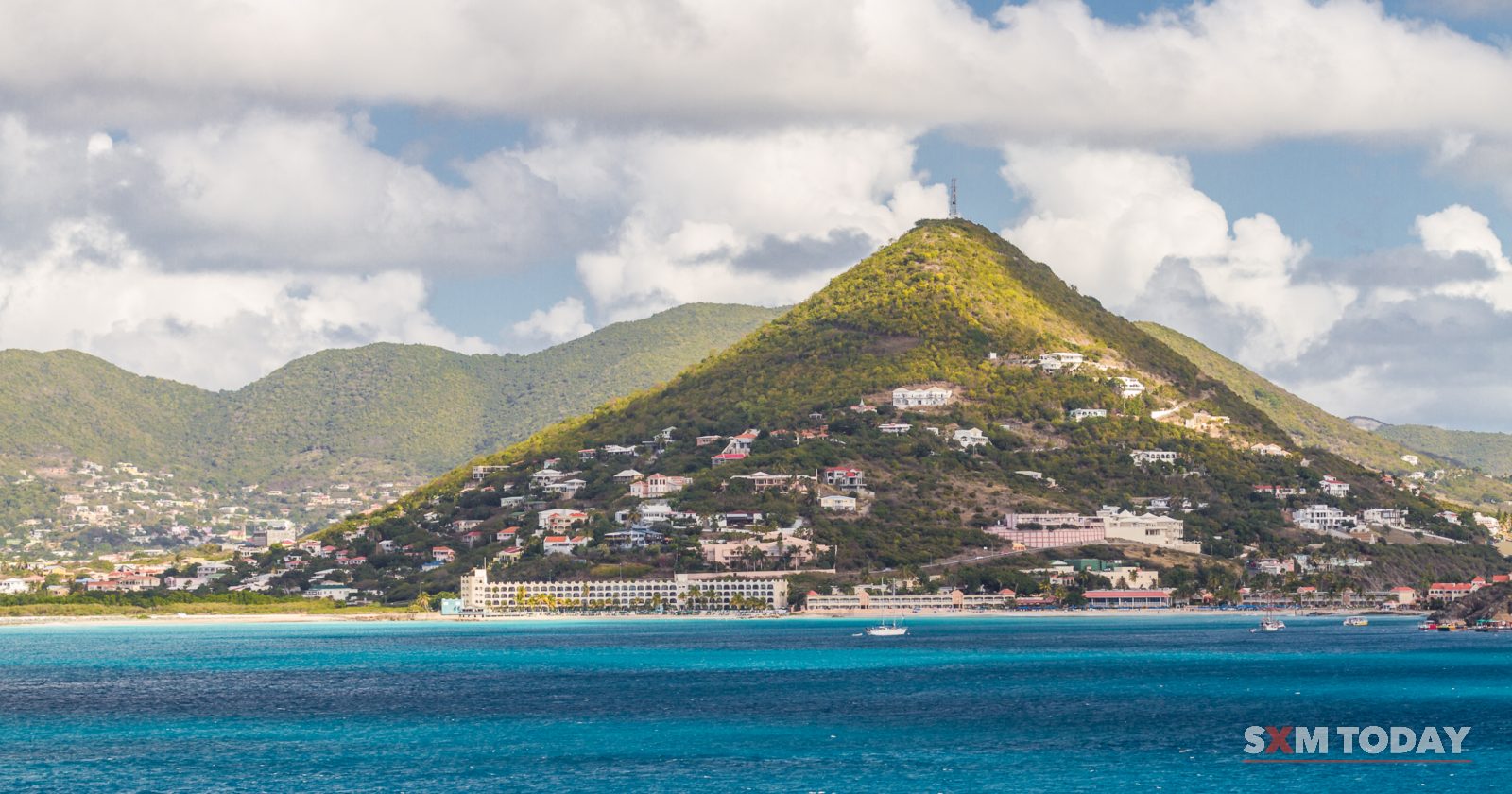 Houses are scattered up a green hill above a strip of buildings and bright blue sea.