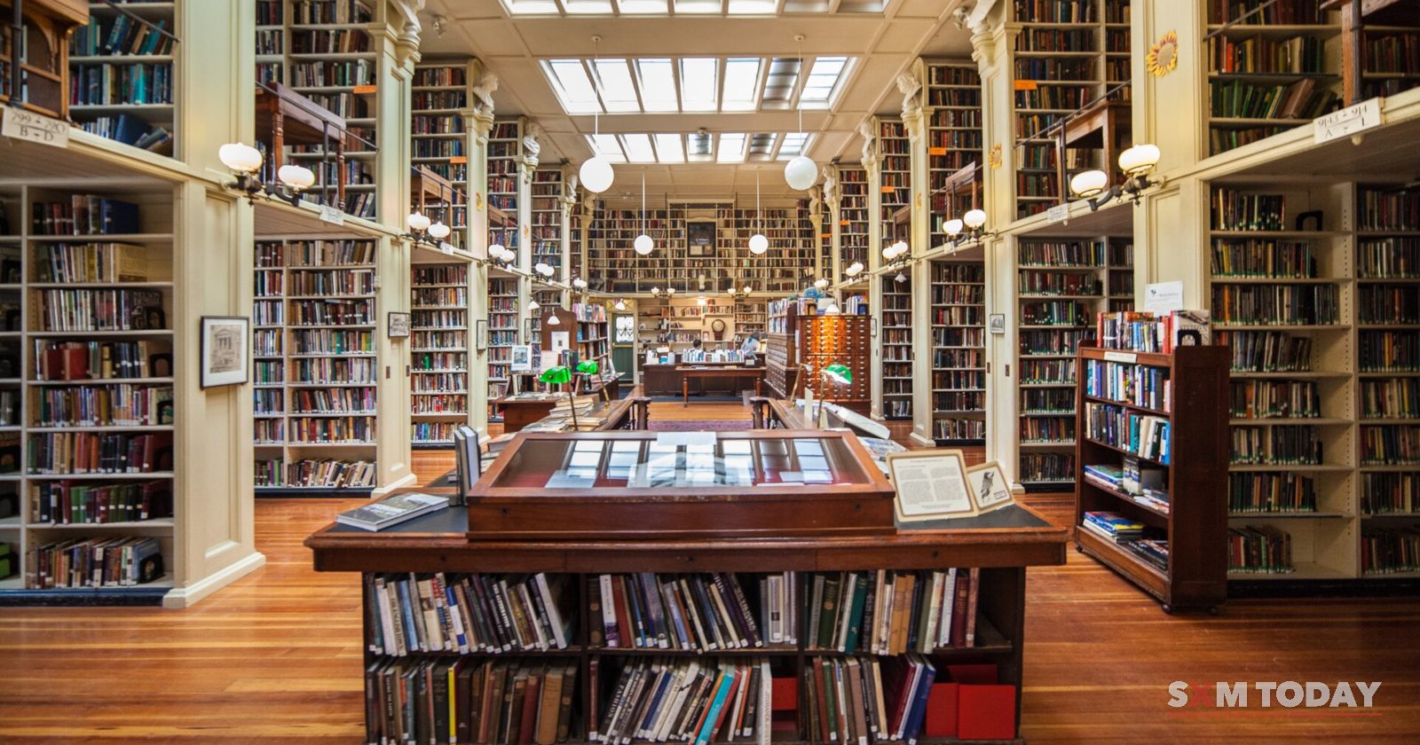 A long library hall lined with wooden bookshelves under skylights, with display cases and tables down the centre.