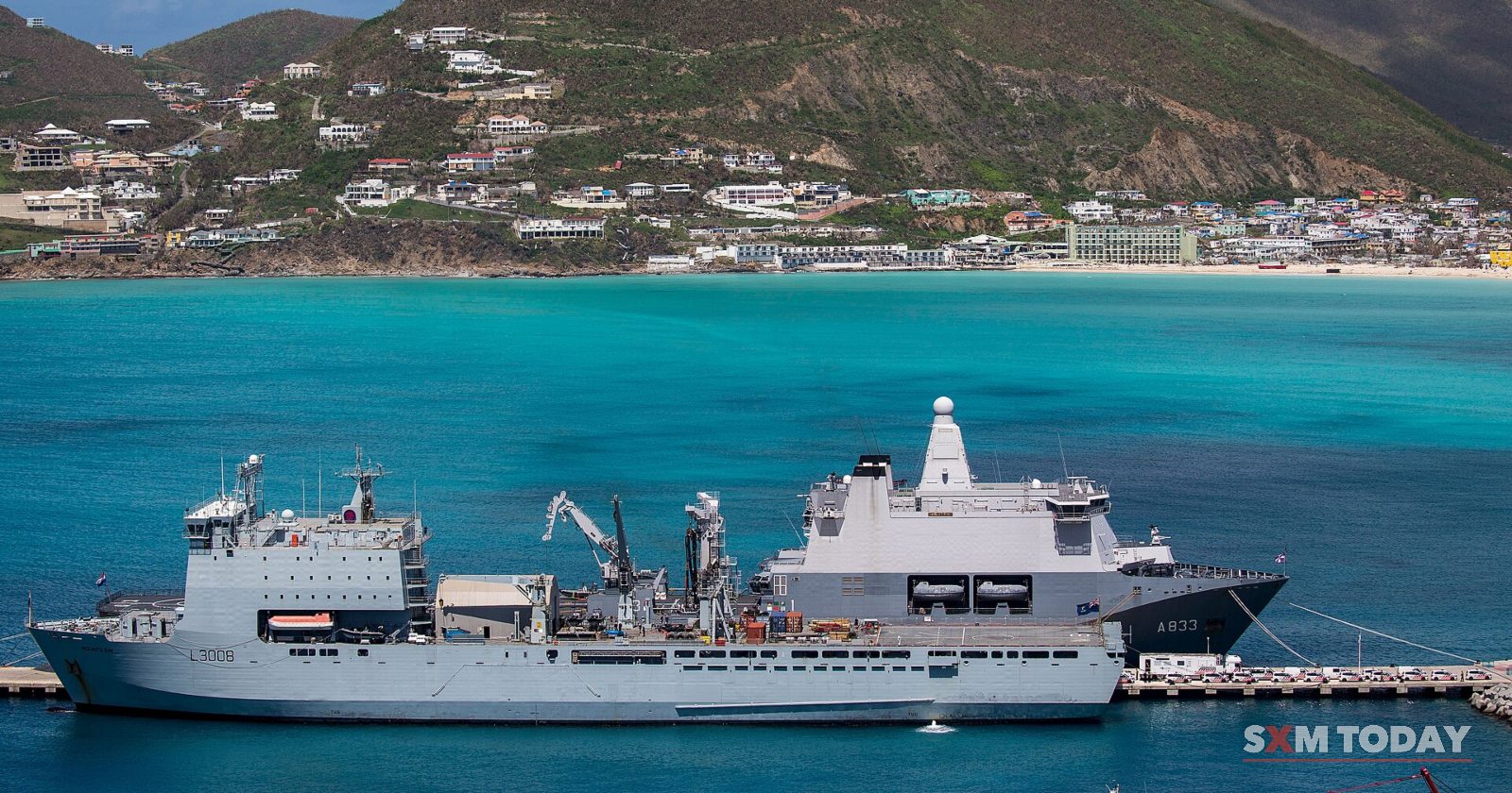 Two grey navy ships are moored at a pier in turquoise water below a hillside of houses.