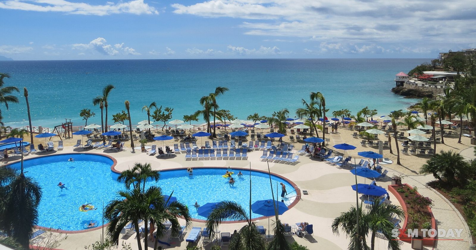 A curved swimming pool ringed by blue loungers and palm trees sits above a beach and turquoise sea under blue sky.