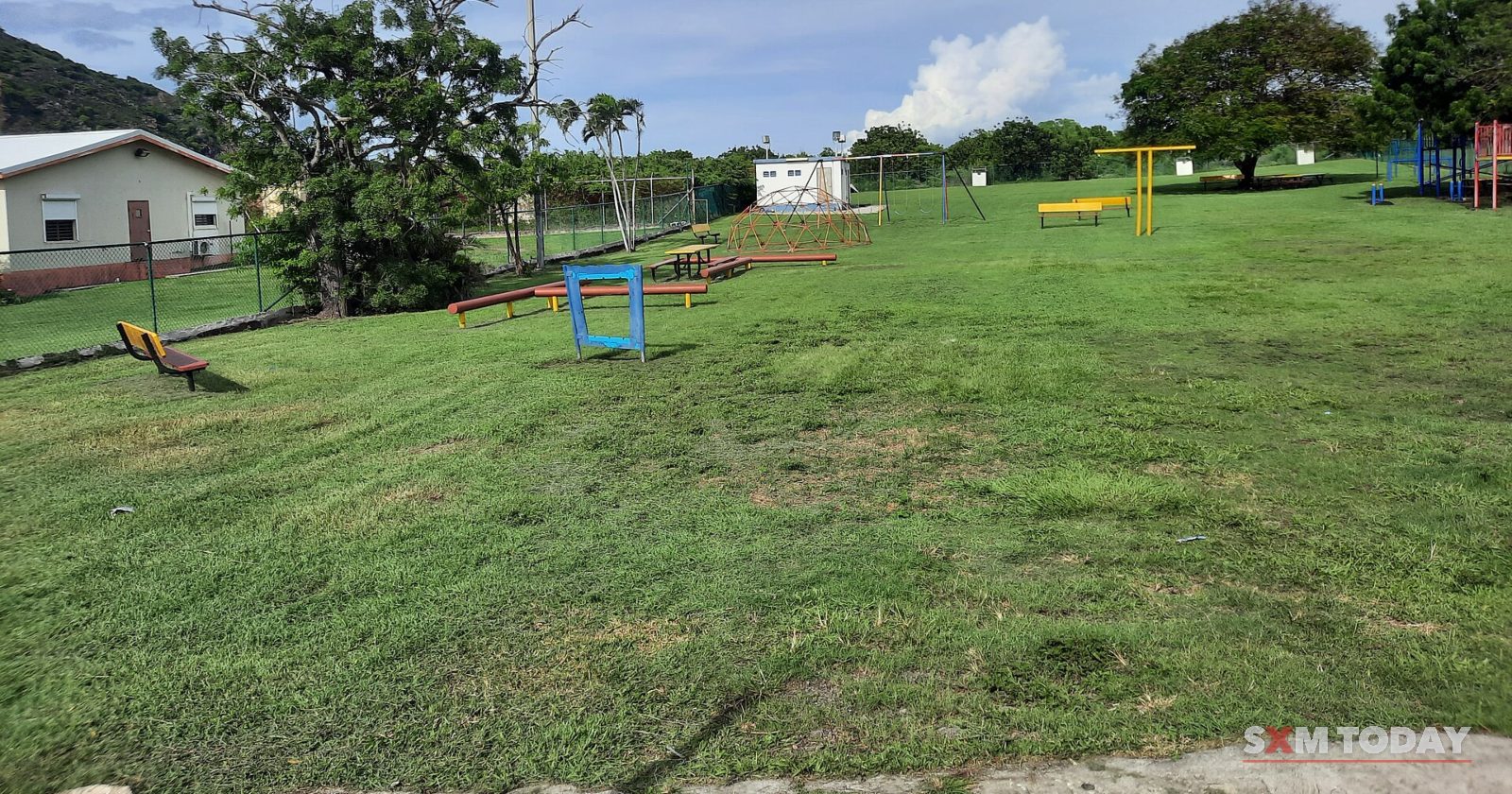 Children's play equipment scattered across a wide green lawn, with trees and a low building on the left.
