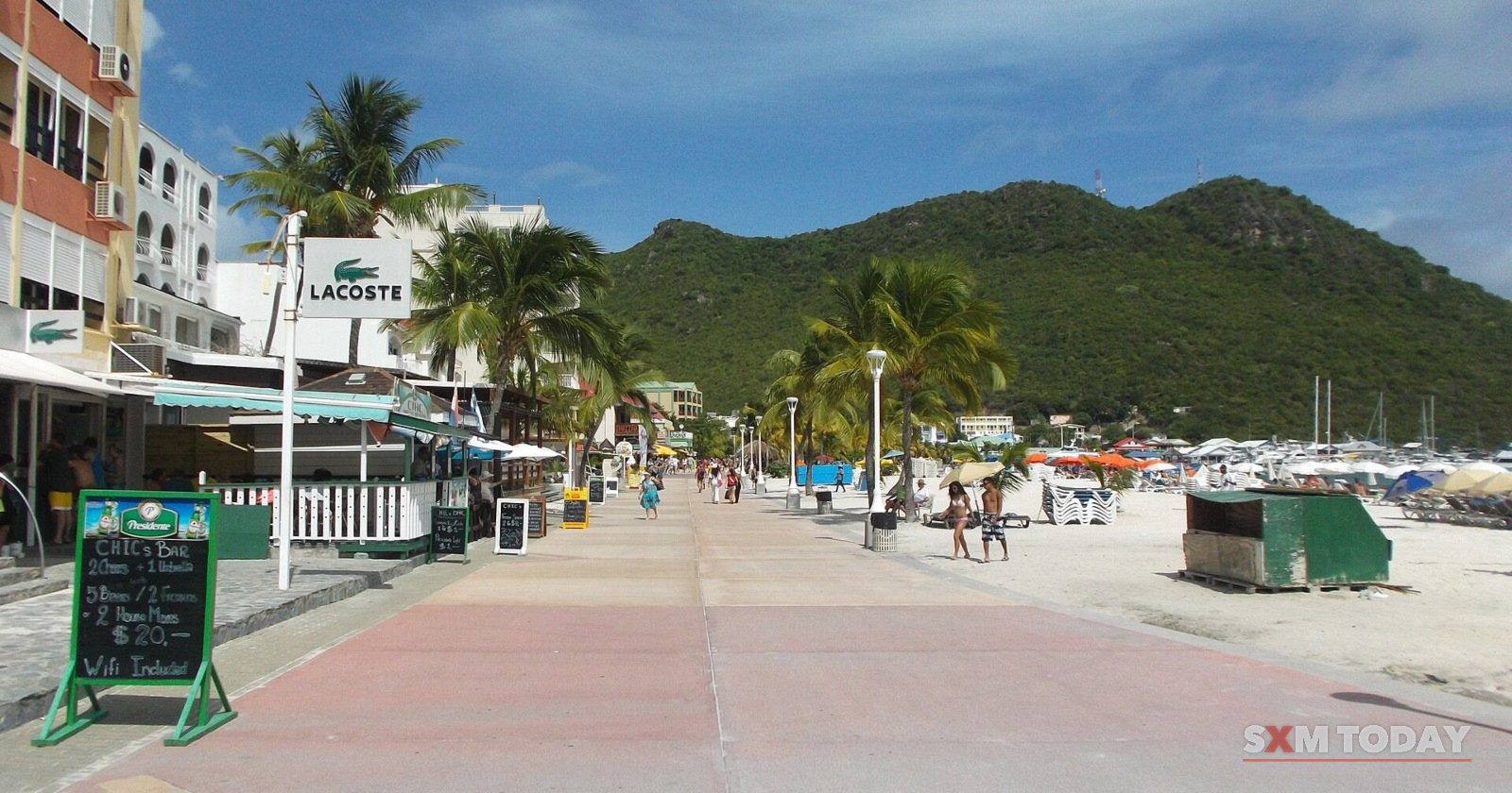 The Great Bay boardwalk in Philipsburg, lined with palm trees, with green hills behind.
