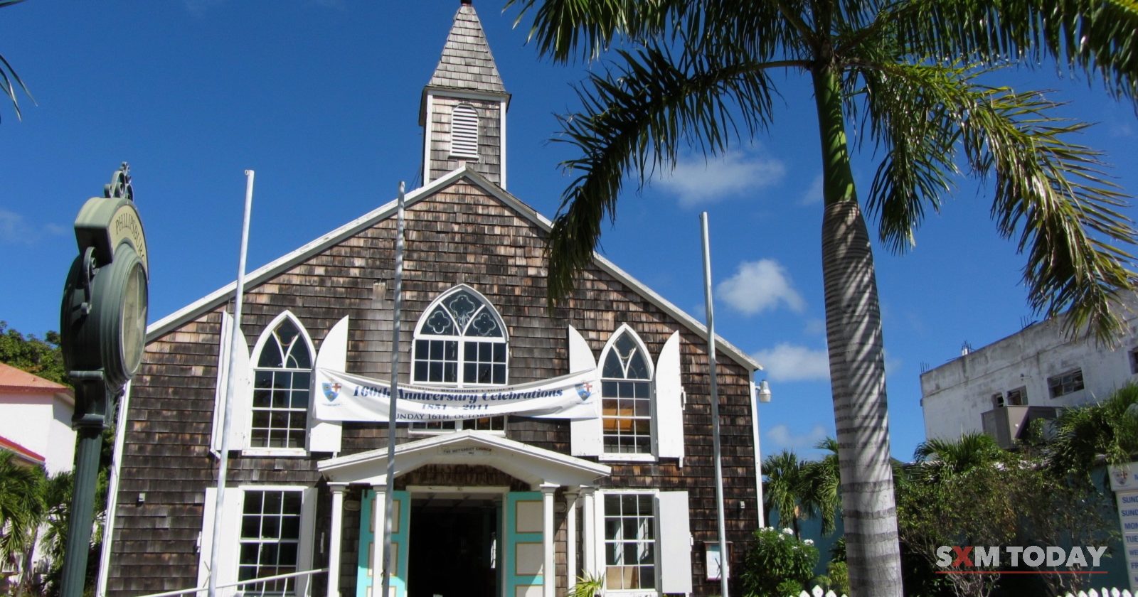 A wooden-shingled church with a white steeple, pointed-arch windows and a white picket fence stands between palm trees under a blue sky.