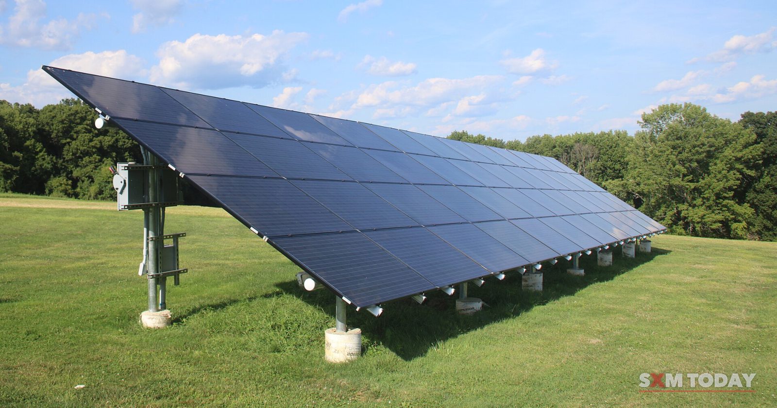 A long tilted bank of solar panels on metal posts stands on a mown lawn in front of trees under a blue sky.
