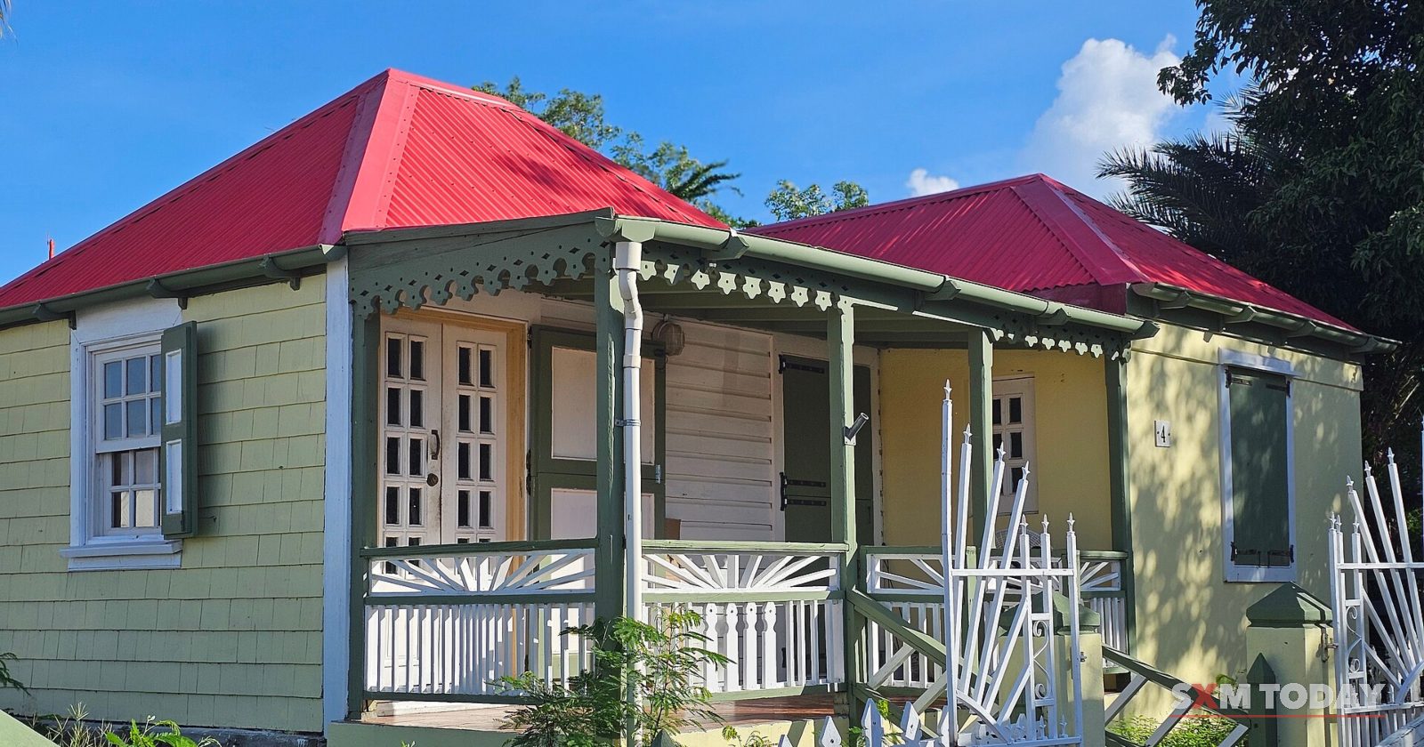 A wooden cottage with green and yellow walls, a red roof and a white fretwork porch behind a white picket fence.