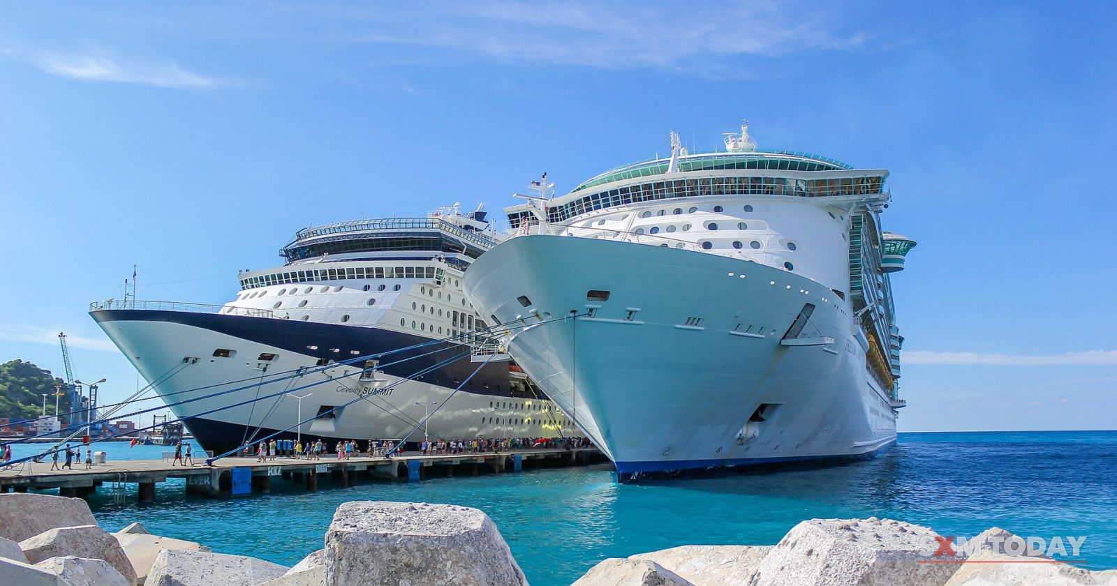 Two large cruise ships are moored side by side at a pier in turquoise water, with concrete breakwater blocks in the foreground.