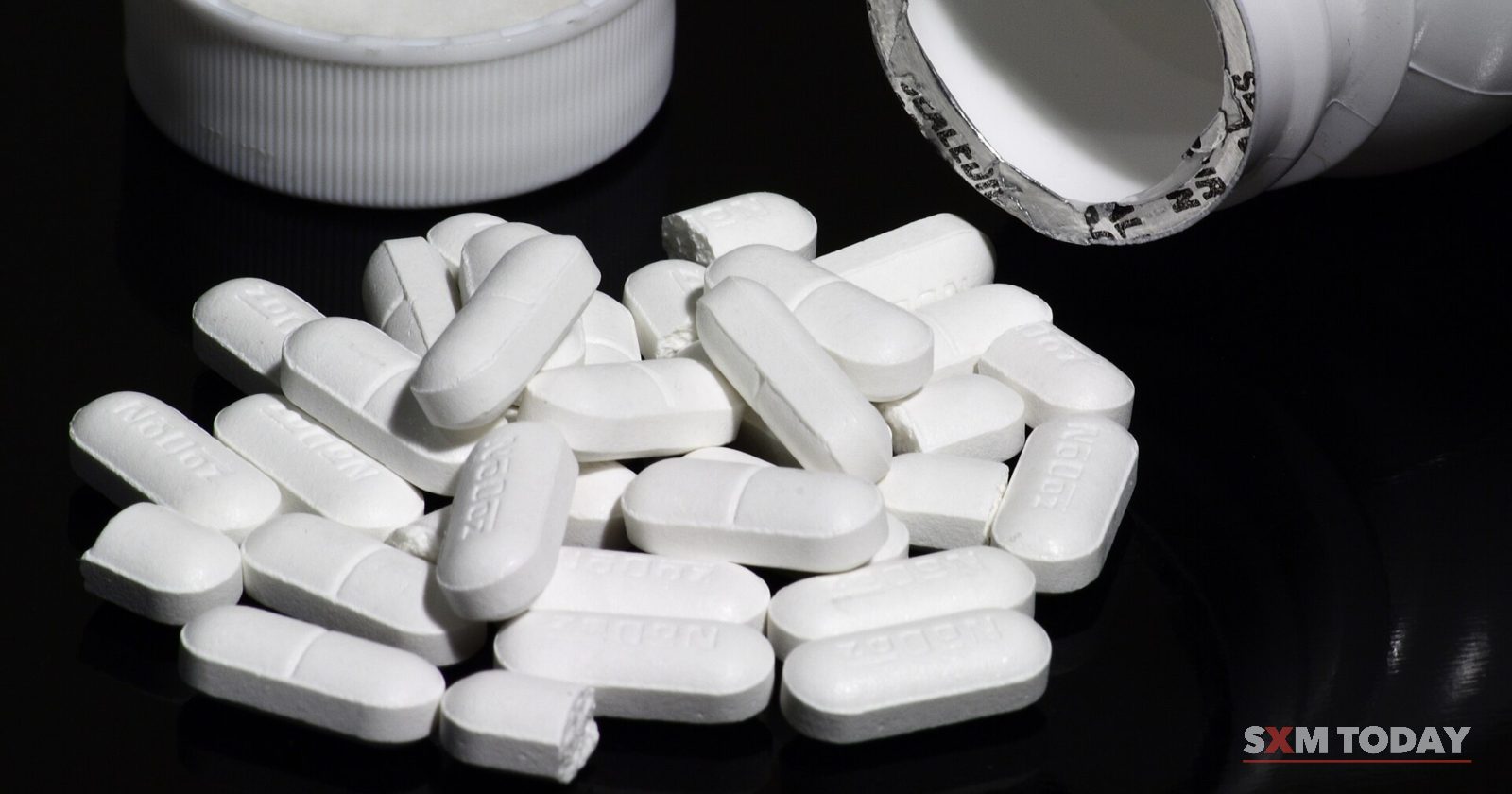 White oblong tablets spilled beside an open white pill bottle and its cap, on a black background.