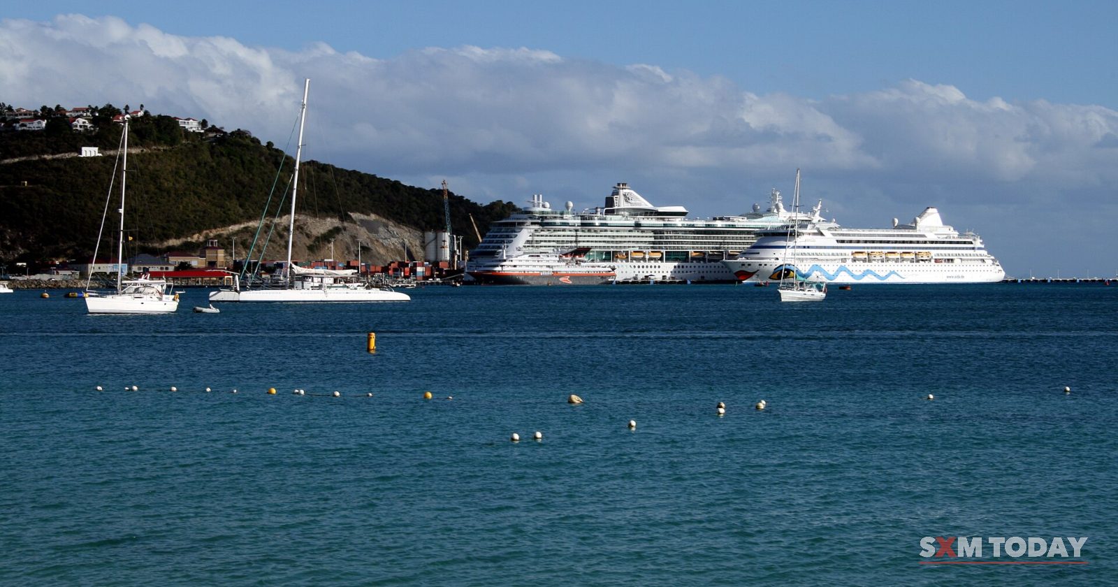 Sailboats ride at anchor on blue water in front of two large white cruise ships below a green headland.