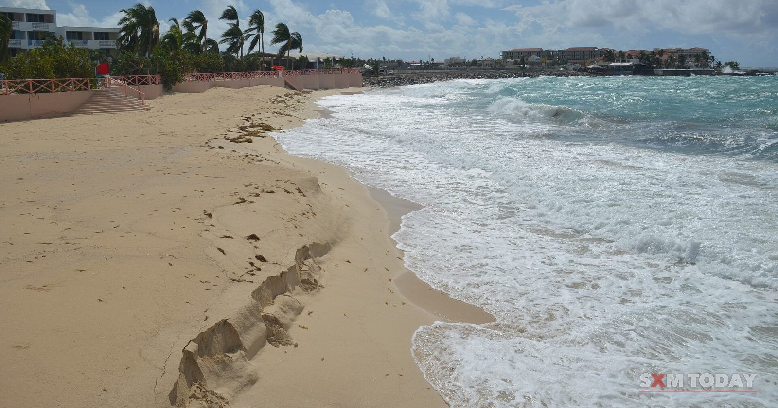 Foaming waves wash over an empty sandy beach as wind bends the palm trees along the shore.