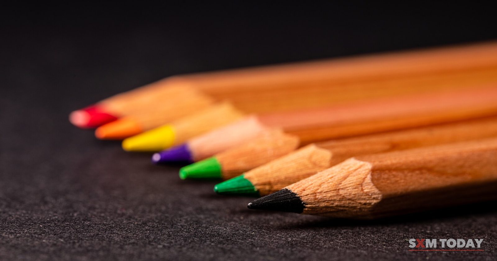 A row of sharpened wooden coloured pencils lies on a dark surface, their tips fanning out in different colours.