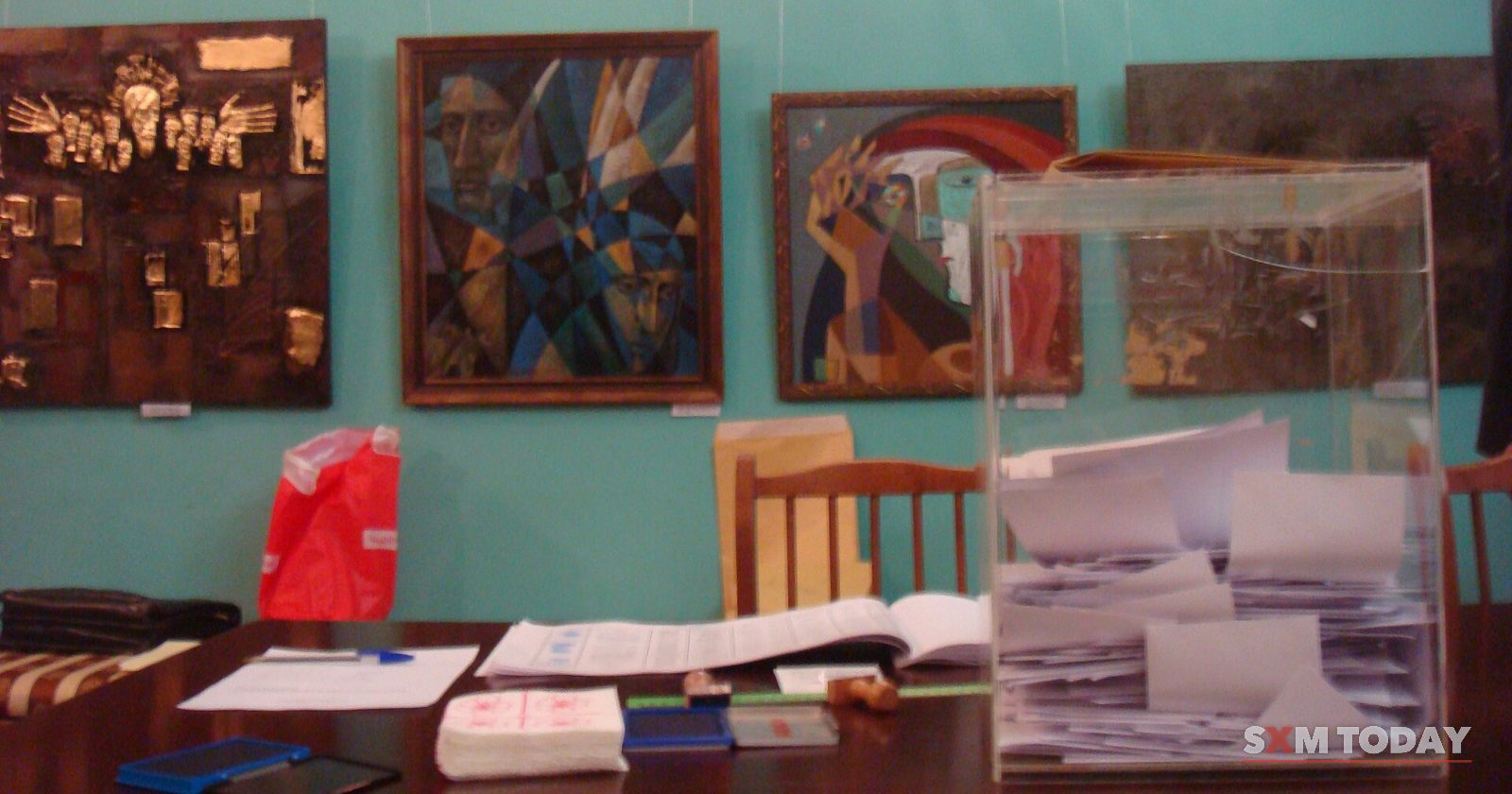 A transparent ballot box half full of folded papers stands on a table in a room hung with paintings.