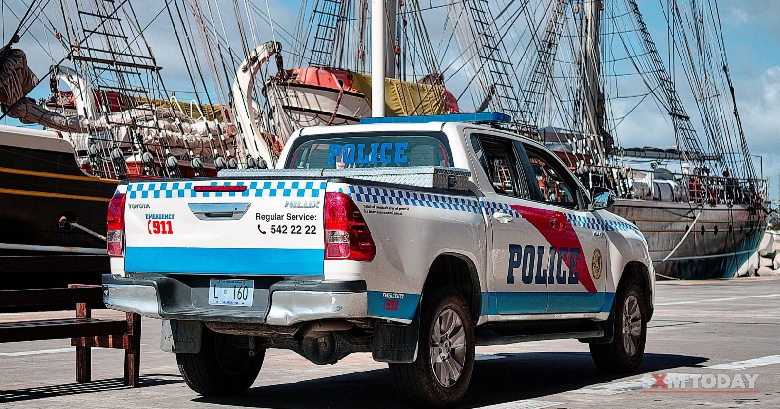 A white Sint Maarten police pickup truck with blue and red markings, parked in front of a tall ship at the harbour.
