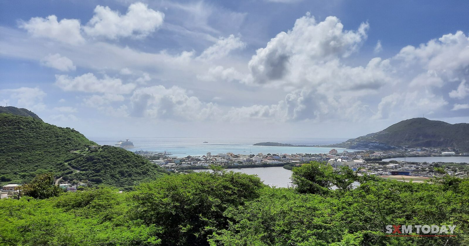From a scrubby hilltop, a view looks across a town and harbour to the sea and distant headlands under a bright, cloudy sky.