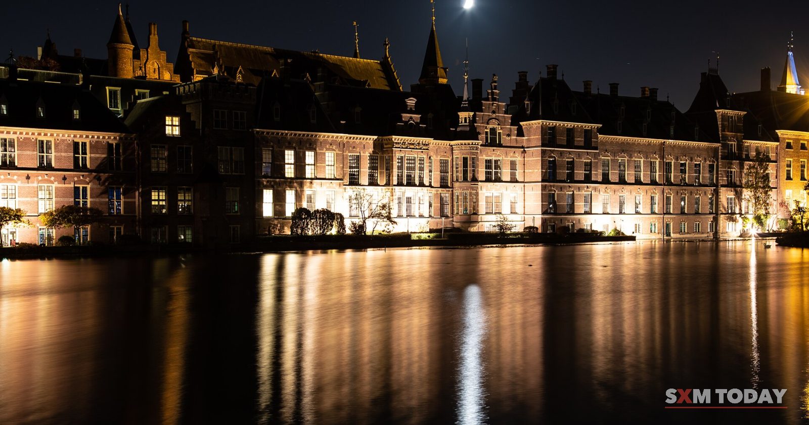A row of lit historic buildings with spires reflected in dark water at night, the moon above.