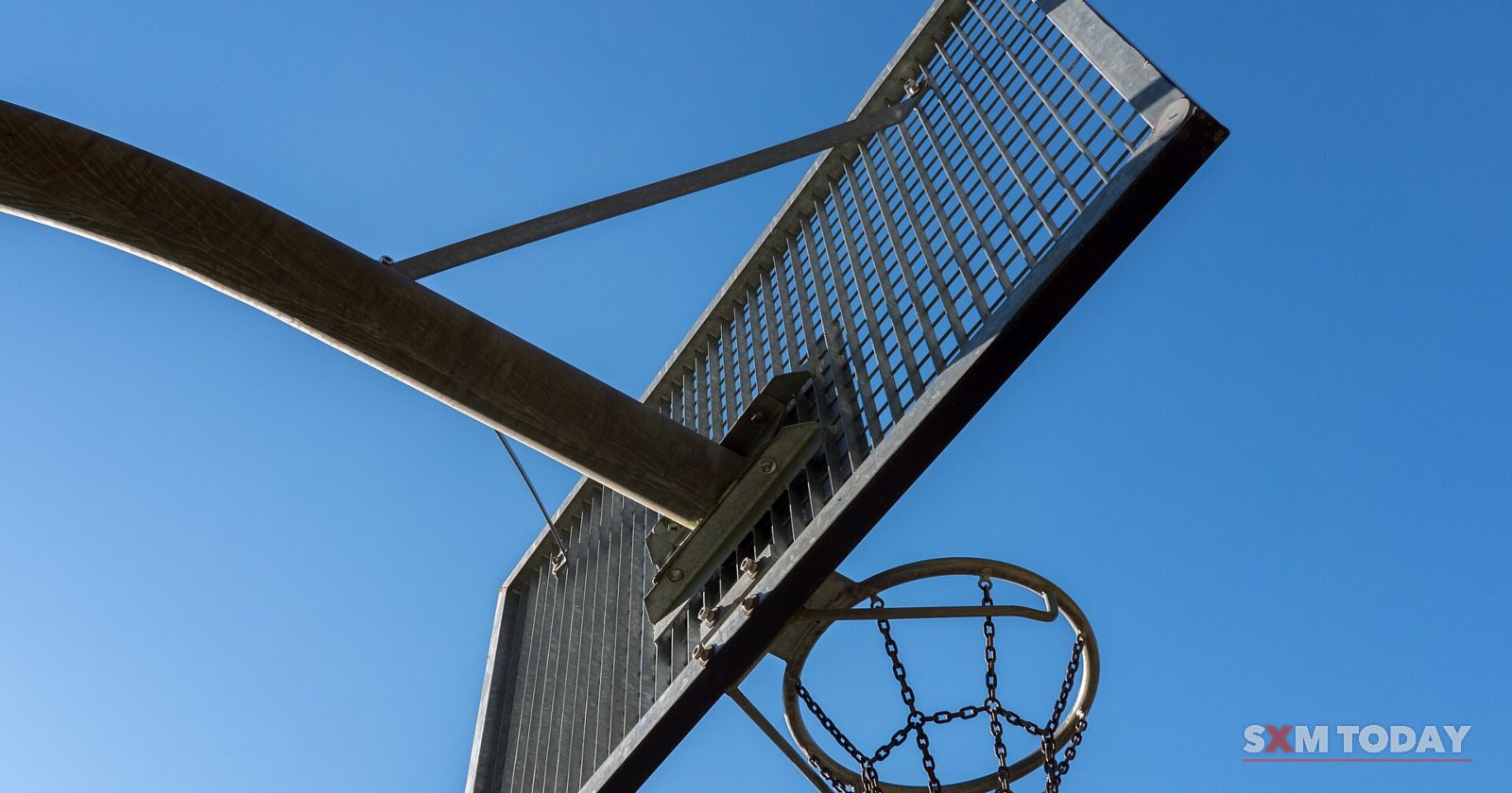 A basketball backboard and hoop with a chain net, seen from below against a clear blue sky.