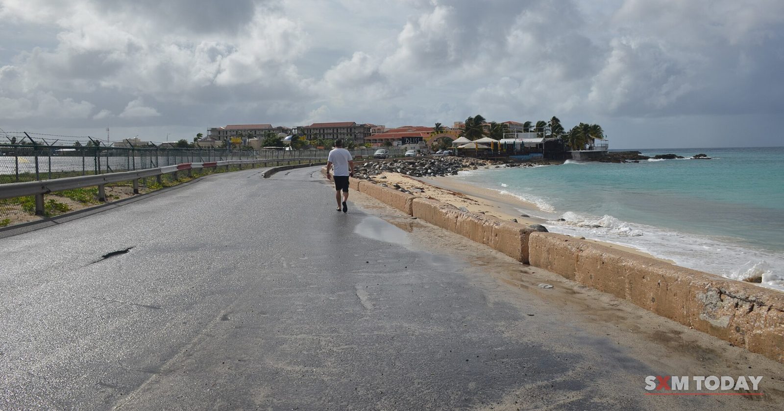 A wet coastal road runs between a chain-link fence and a low sea wall under heavy grey cloud, with one person walking in the distance.