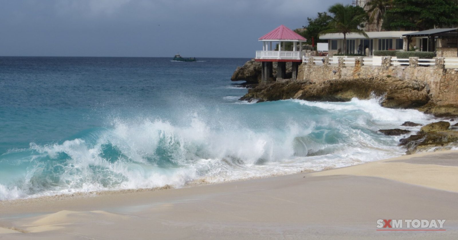 A large wave breaks onto a sandy beach below a rocky point with a pink-roofed gazebo and buildings among trees.