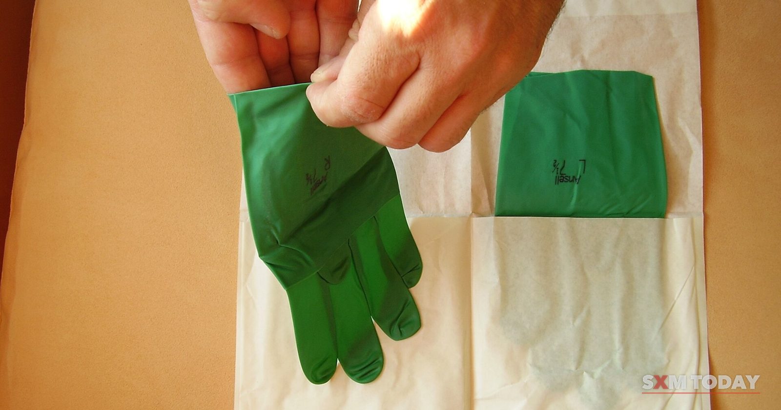 Hands lifting a green surgical glove out of its opened sterile paper wrapper, with the second glove still inside.