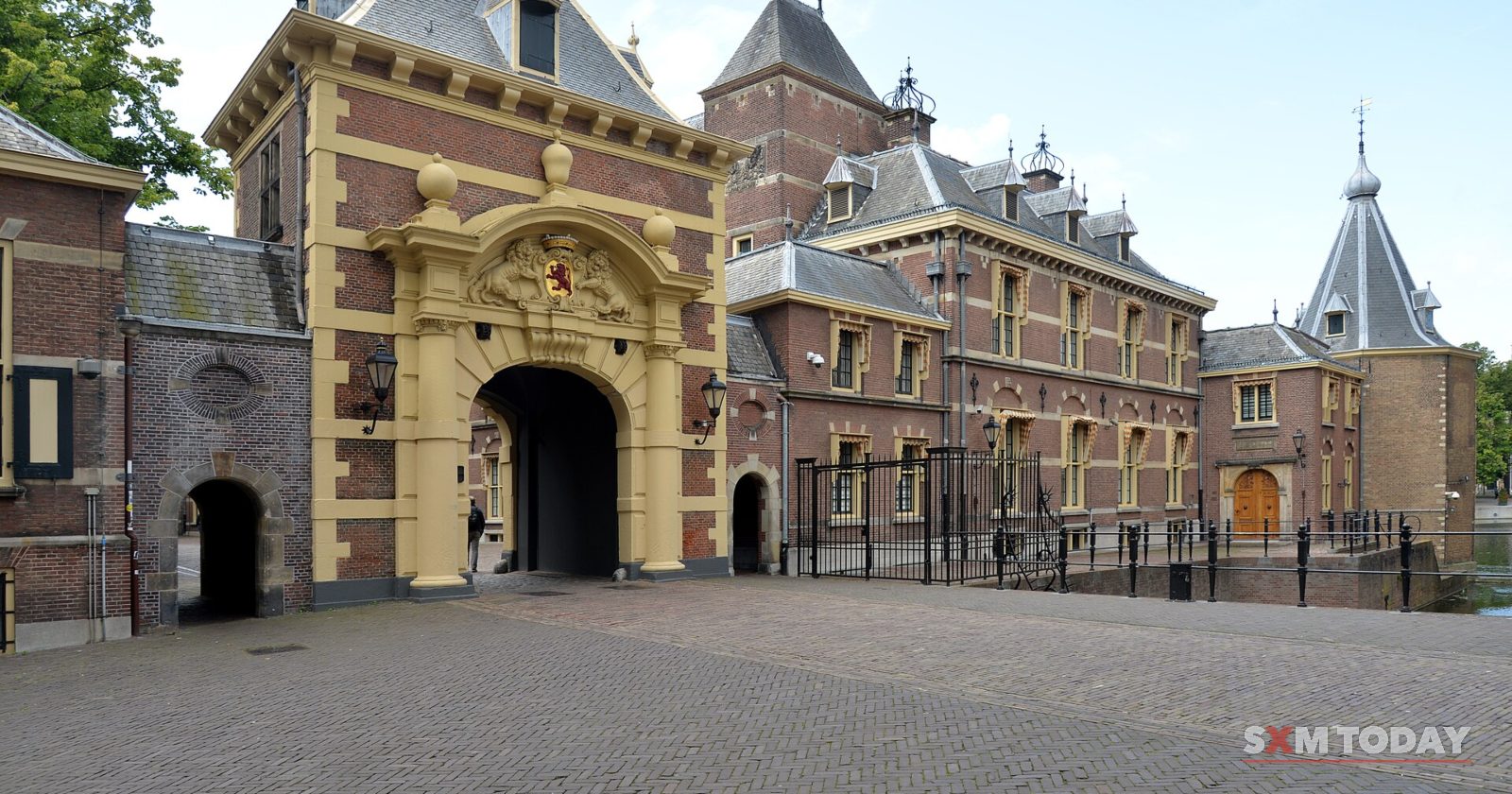 An ornate sandstone gateway with a coat of arms between brick buildings, facing an empty cobbled square.
