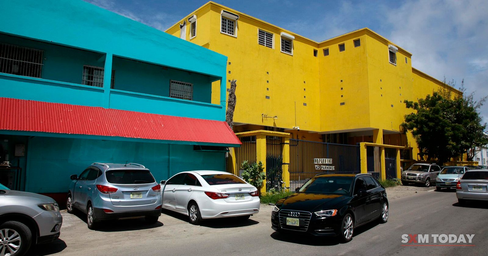 Turquoise and yellow buildings stand beside a car park with several parked cars under a blue sky.