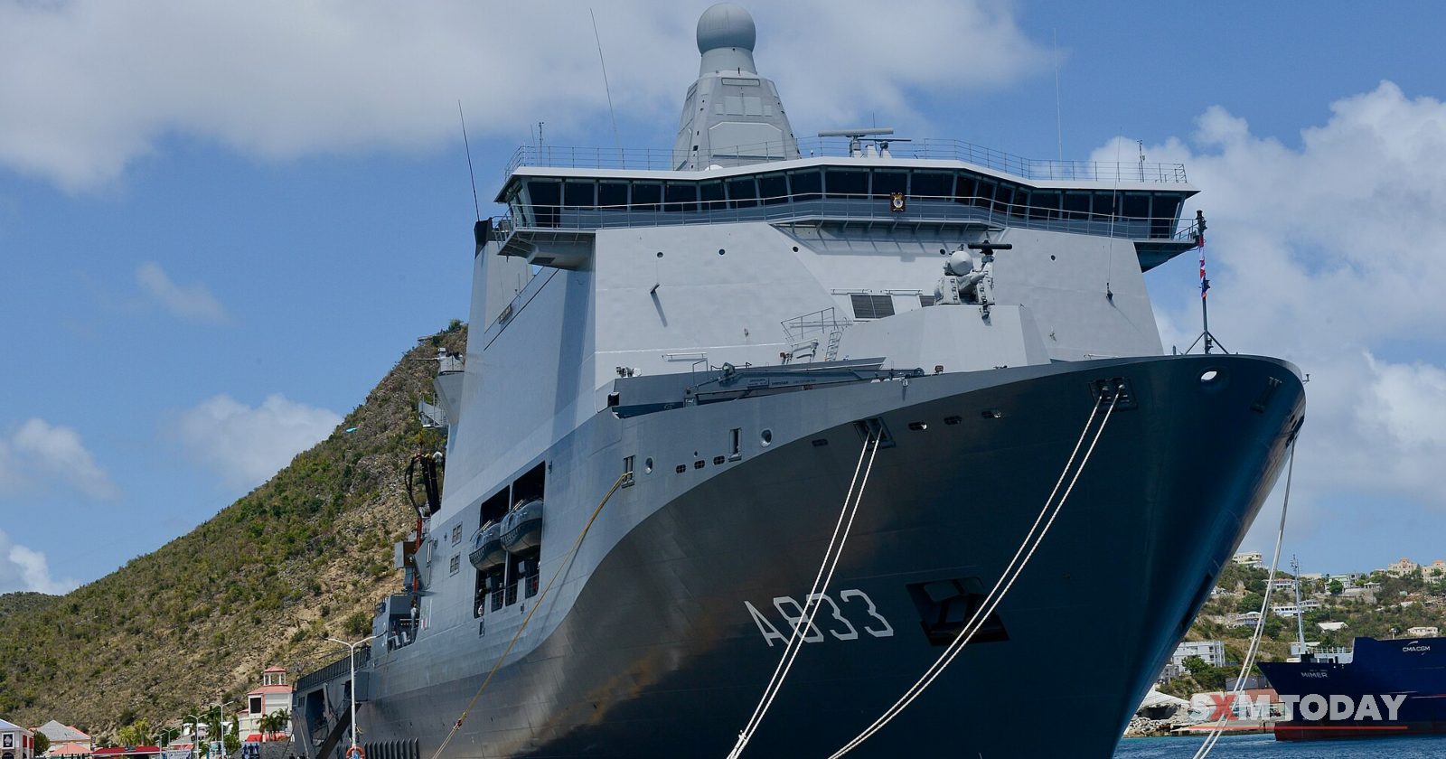 A grey naval support ship marked A833 is moored at a pier below a green hill.