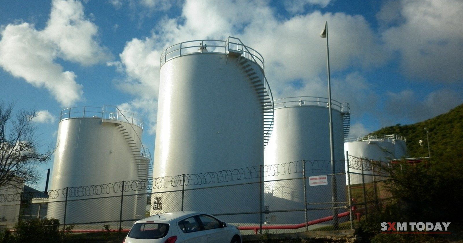 Three white fuel storage tanks behind a fence, with a small car parked in front.