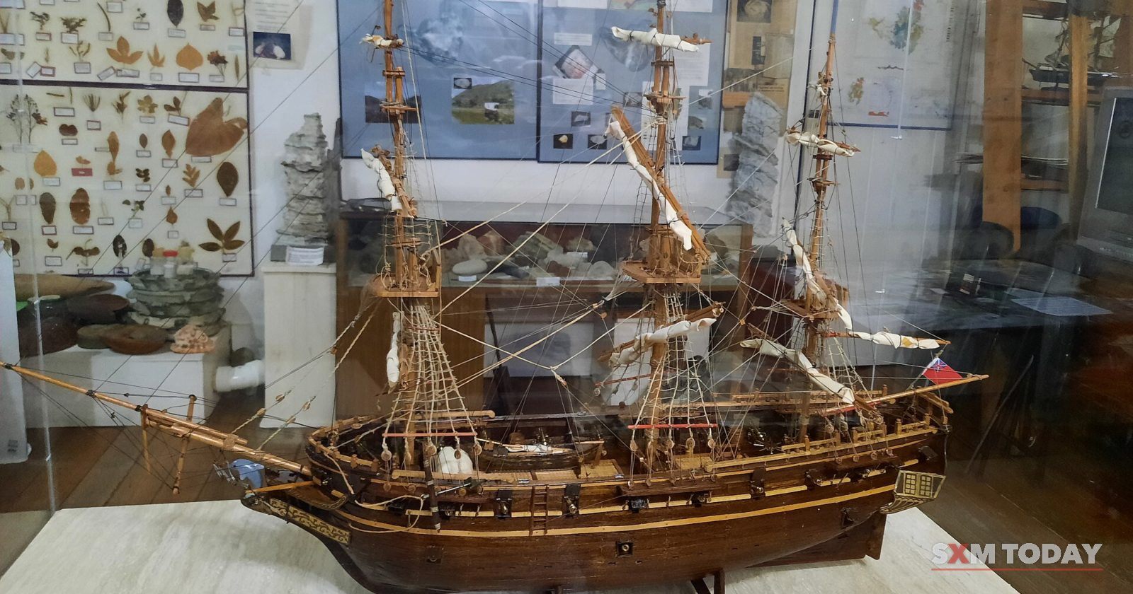 A detailed wooden model of a three-masted sailing ship sits in a glass case in a museum room.