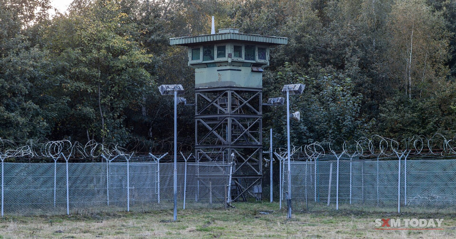 A guard tower on a steel frame stands behind a double chain-link fence topped with razor wire, with floodlights in front of it.