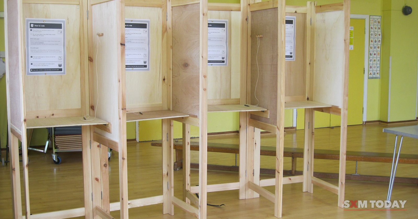 A row of empty wooden polling booths with voting instructions pinned inside, in a hall with green walls and a wooden floor.