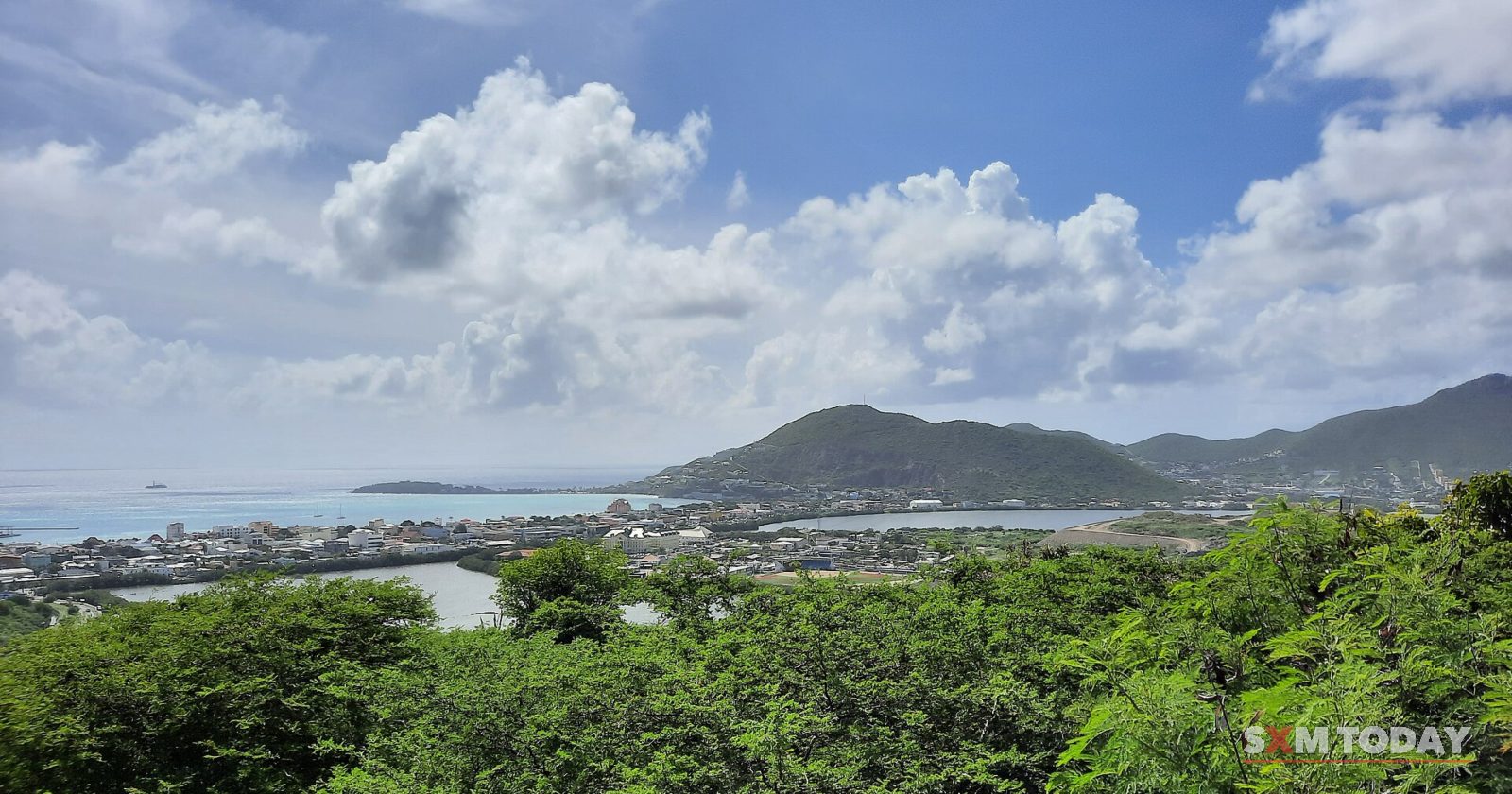 Green scrub in the foreground gives way to a view of a low town, a salt pond and a bay ringed by hills under white clouds.