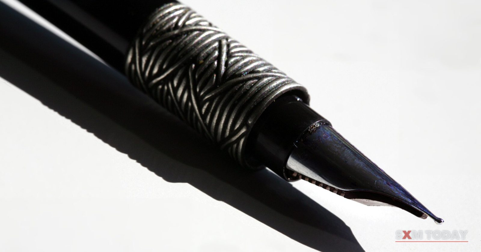 Close-up of a black fountain pen nib with an engraved silver band, casting a sharp shadow on a white surface.