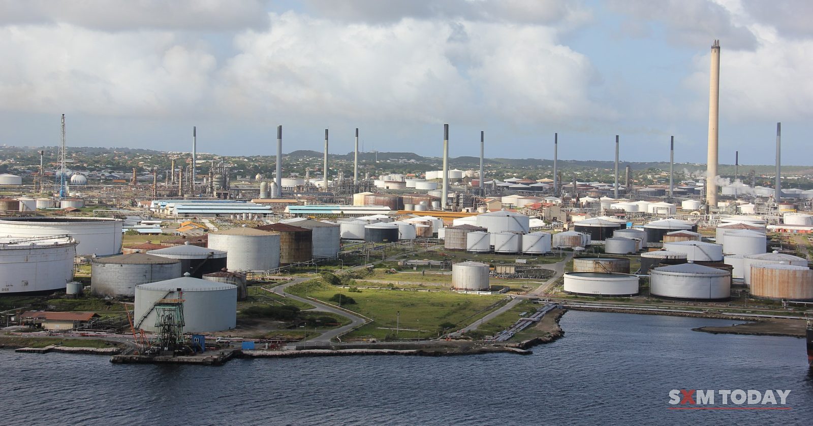 Rows of white storage tanks and chimneys of an oil refinery stretch along the waterfront under a cloudy sky.