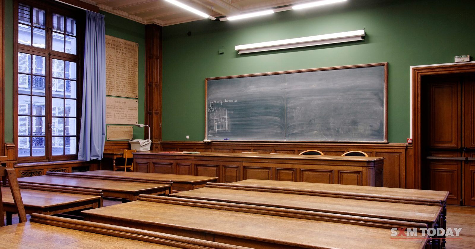 An empty classroom with rows of wooden desks facing a blackboard under fluorescent lights.