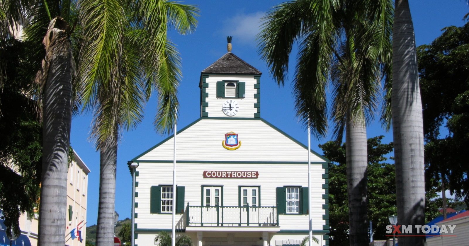 A white colonial courthouse with a green-trimmed cupola and clock stands between tall royal palms under a deep blue sky.