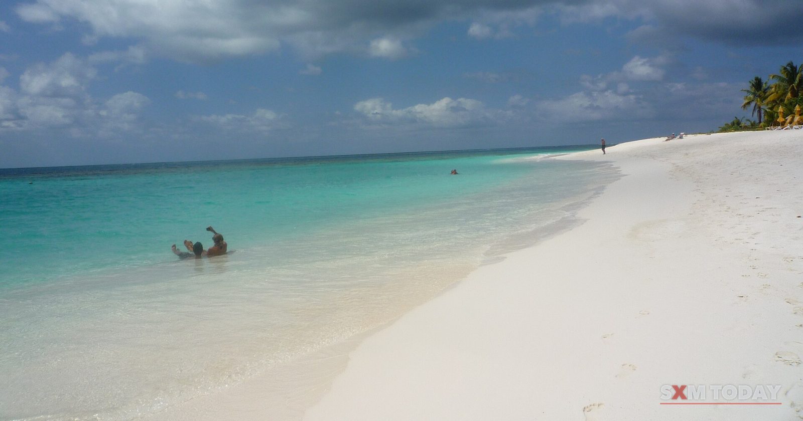 A long curve of white sand beach meets shallow turquoise water under a cloudy sky, with a lone distant swimmer.