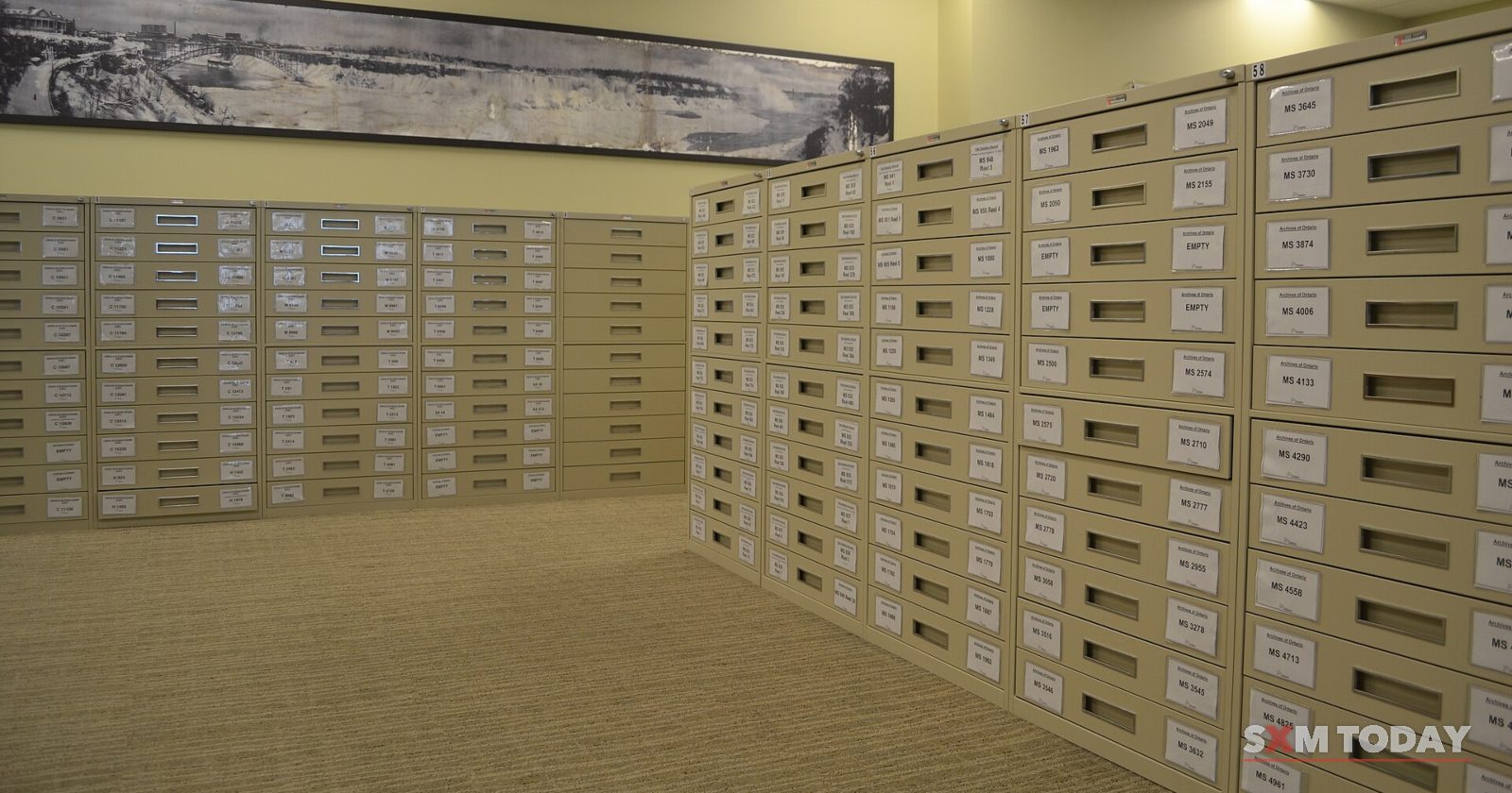 Long banks of pale filing drawers with small labels line a carpeted archive room under ceiling lights.