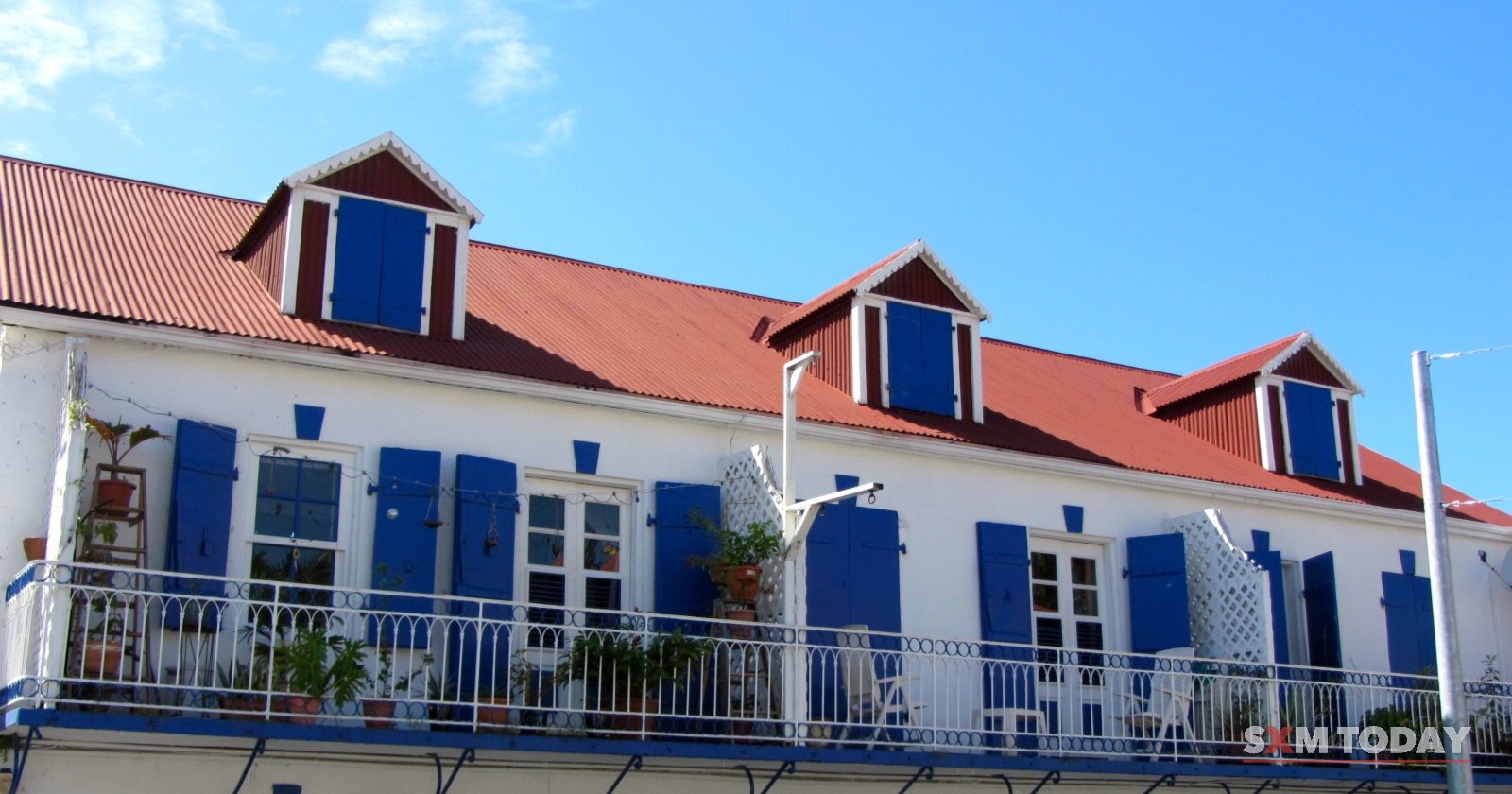 A white two-storey building with blue shutters, dormer windows and a red roof stands against a clear sky.