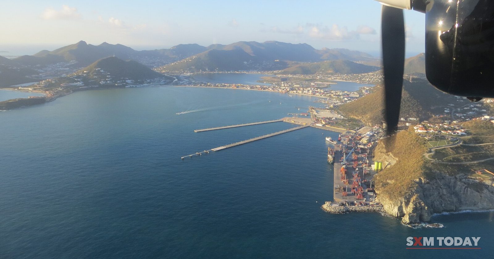 An aerial view from an aircraft window, with a propeller at the edge, looks down on a wide blue bay, its piers and a container terminal below green hills.